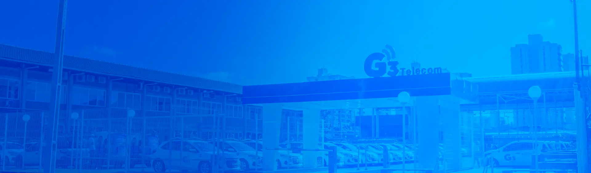 Foto da fachada da sede da G3 Telecom em Teresina, Piauí. A imagem, com um filtro azul, exibe o prédio comercial do provedor de internet, com o letreiro da empresa na entrada e os carros da frota estacionados em frente.